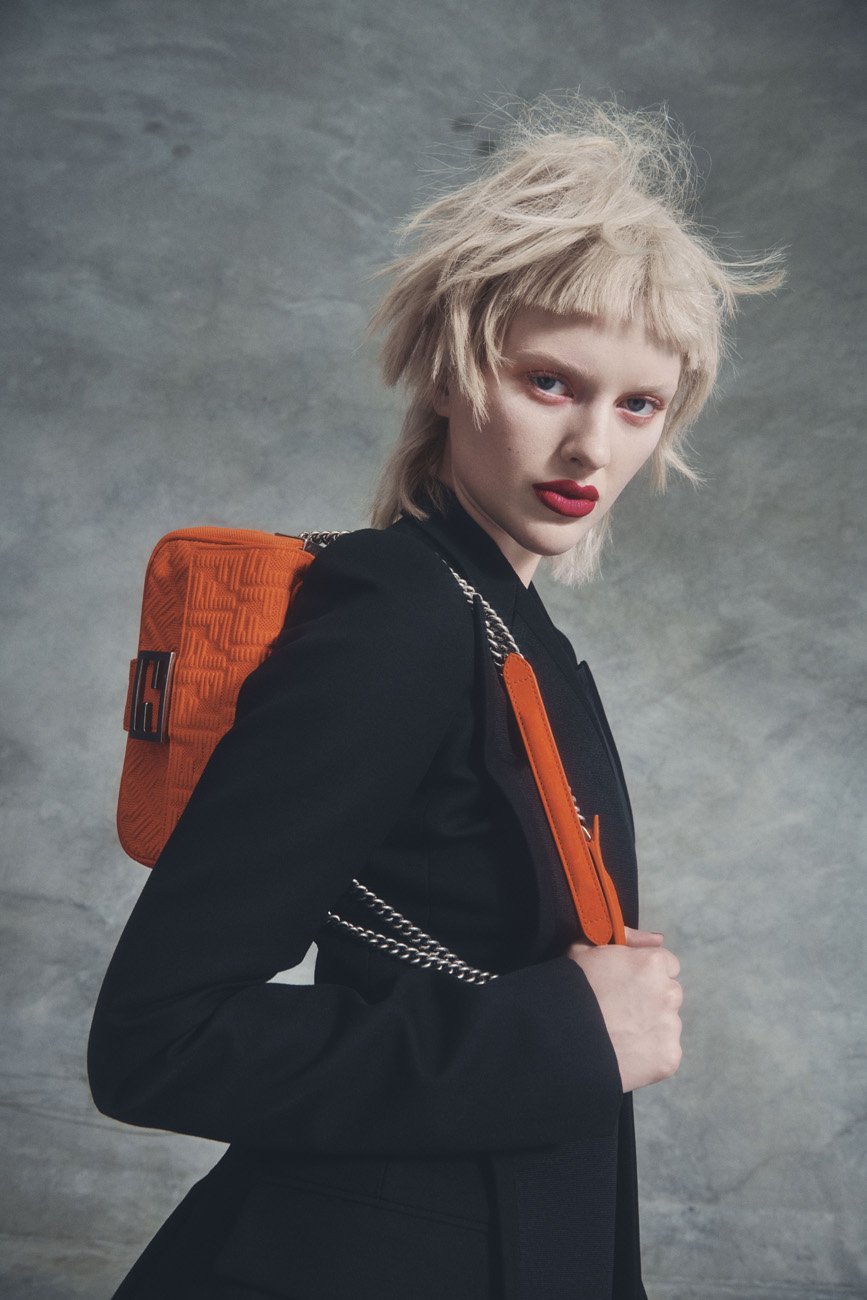 Fashion portrait of a woman with a blonde mullet hairstyle, wearing a black jacket and carrying an orange corduroy bag with a silver chain.