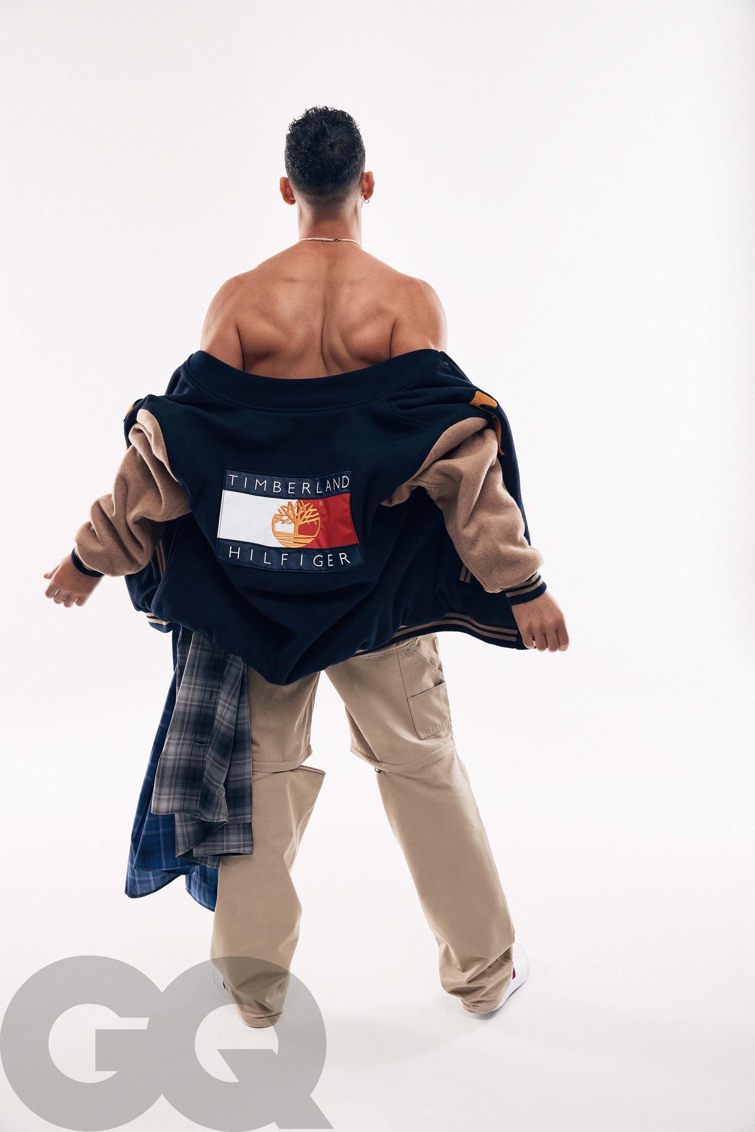 Shirtless adult male modeling a blue and tan Timberland Hilfiger varsity jacket and khaki pants in a fashion studio shot on white.