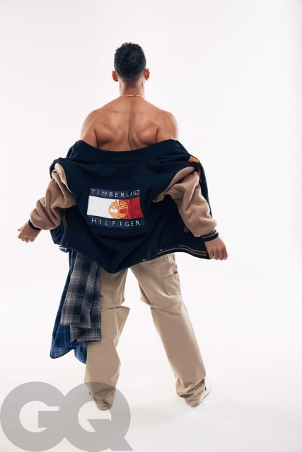 Shirtless adult male modeling a blue and tan Timberland Hilfiger varsity jacket and khaki pants in a fashion studio shot on white.