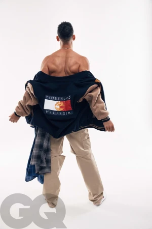 Shirtless adult male modeling a blue and tan Timberland Hilfiger varsity jacket and khaki pants in a fashion studio shot on white.