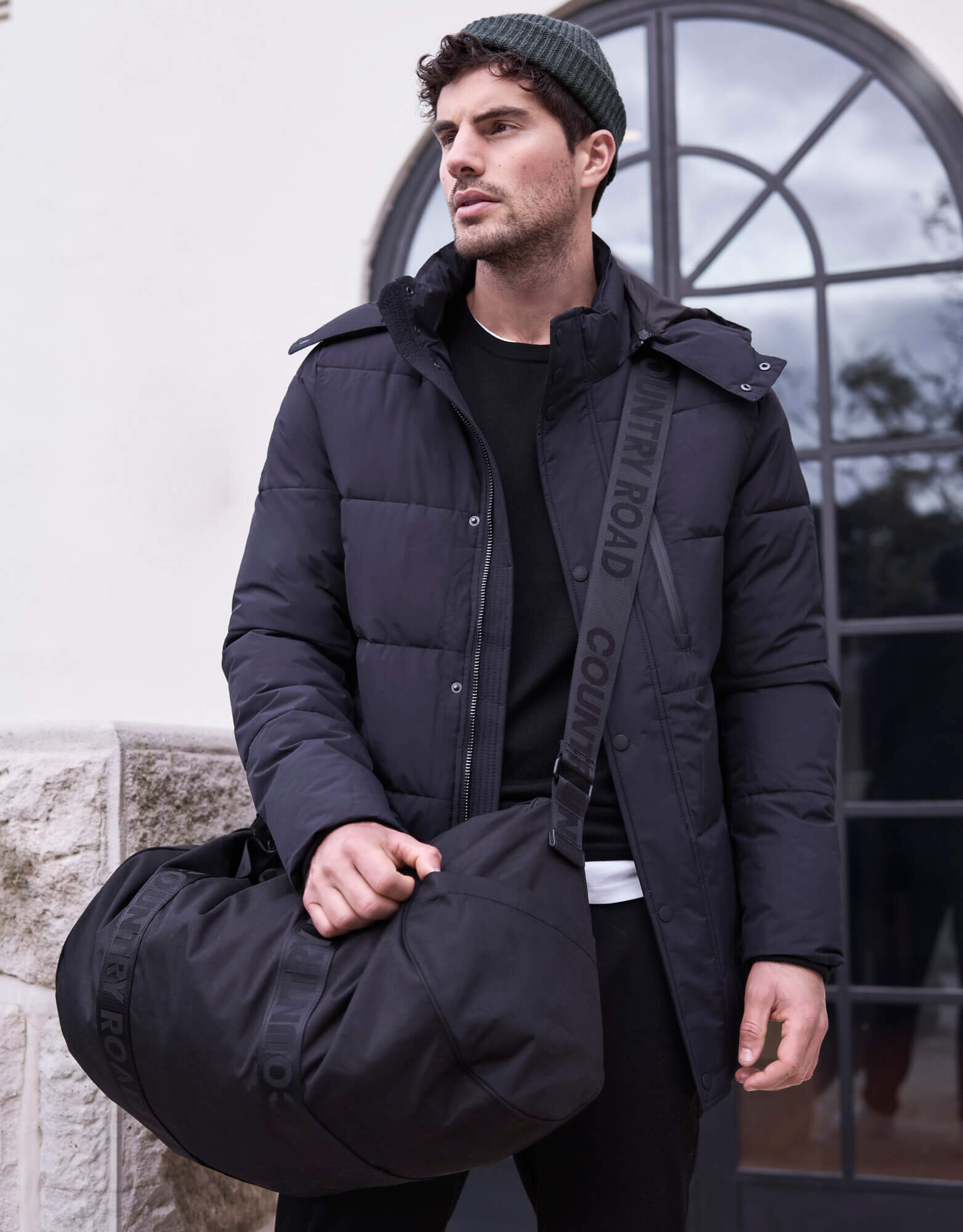 A man in a puffer jacket and beanie stands outside with a black duffel bag slung over his shoulder in an outdoor, urban setting.