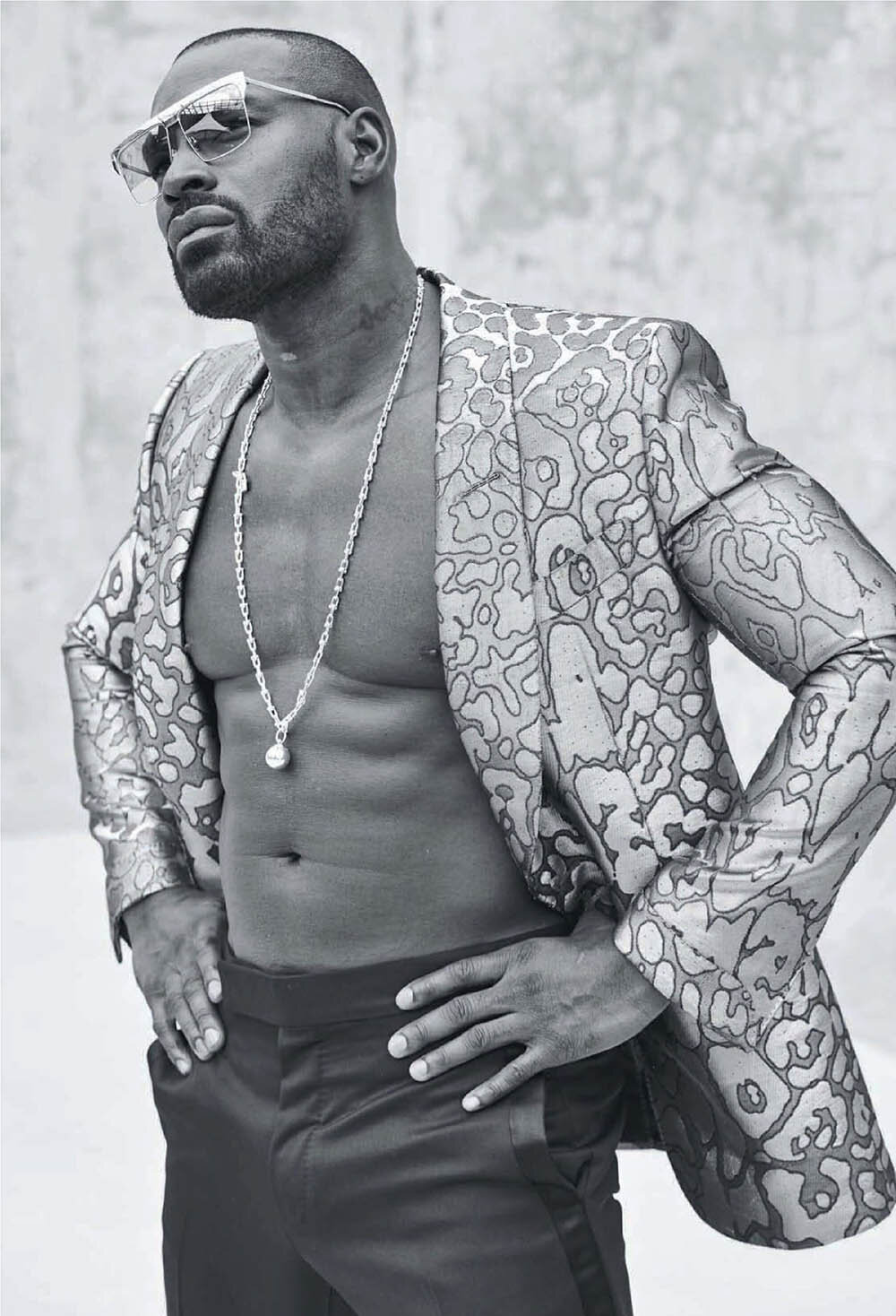 Black male model in a floral suit and sunglasses strikes a confident pose against a grey background, showcasing style and physique.