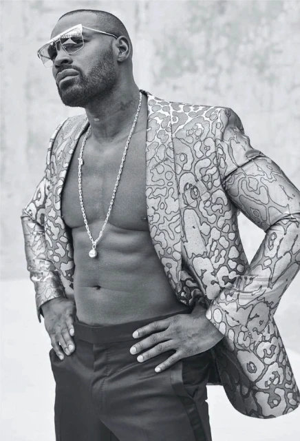 Black male model in a floral suit and sunglasses strikes a confident pose against a grey background, showcasing style and physique.