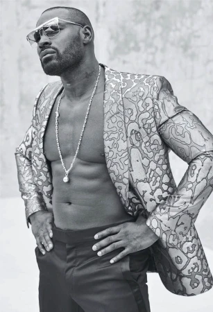 Black male model in a floral suit and sunglasses strikes a confident pose against a grey background, showcasing style and physique.