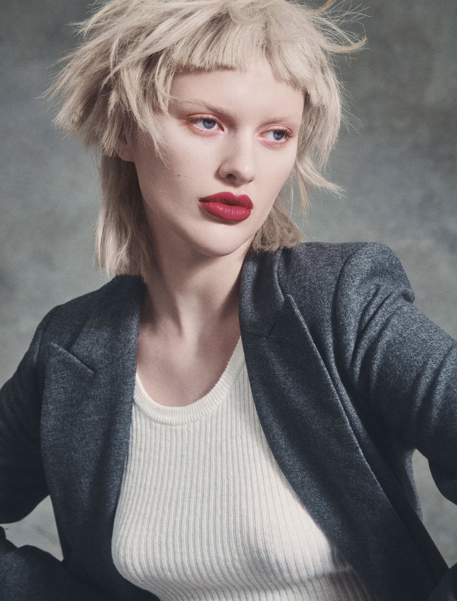 Portrait of a fair-skinned woman with blonde hair, wearing a grey blazer and a ribbed white top against a grey background.