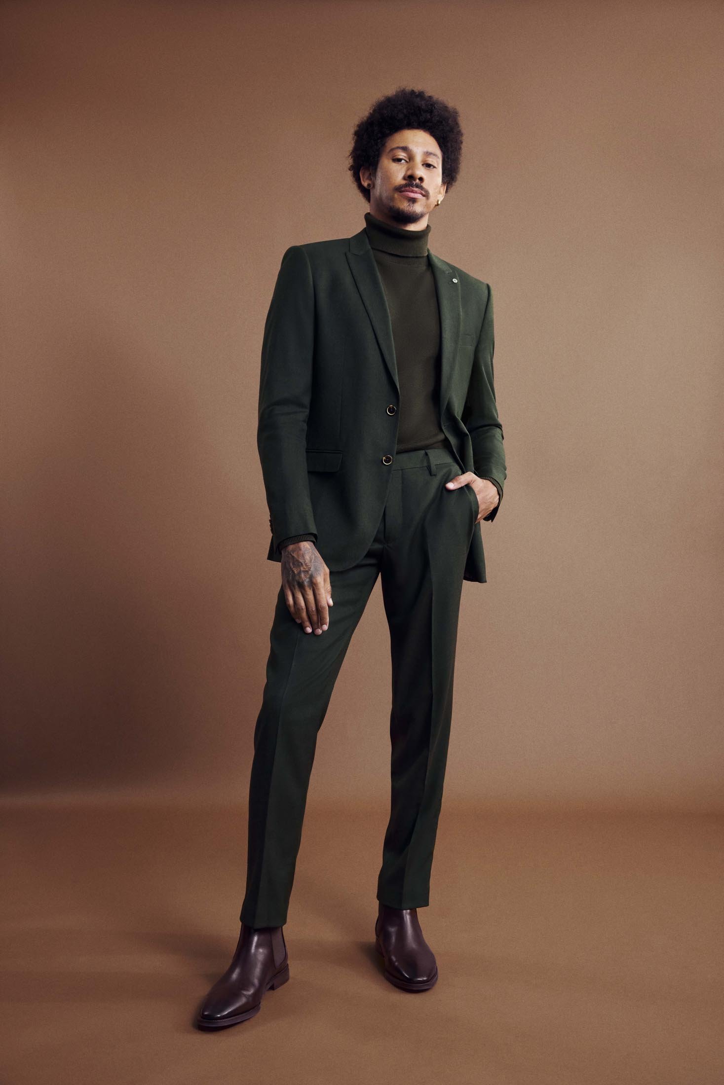 A stylish adult male with an afro wearing a green suit stands confidently against a brown backdrop in a fashion portrait.