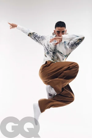 A man captured mid-air in a dynamic dance jump wearing a printed hoodie and brown trousers against white seamless background
