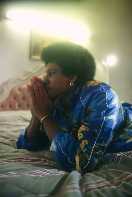 Woman with afro laying on striped bed in patterned blue jacket. Close-up shot, bedroom setting, indoor lighting, soft focus, contemplative mood.