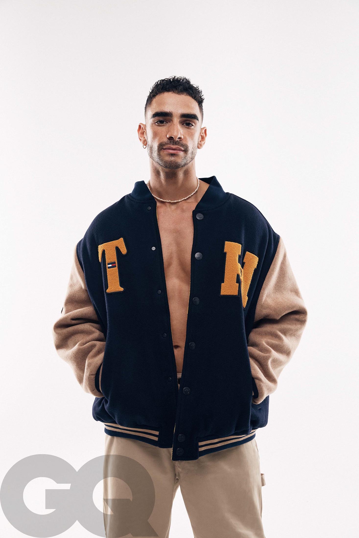 Portrait of a fit, light-skinned male wearing a varsity jacket with open front, neutral pants, and gold chain against a white backdrop.