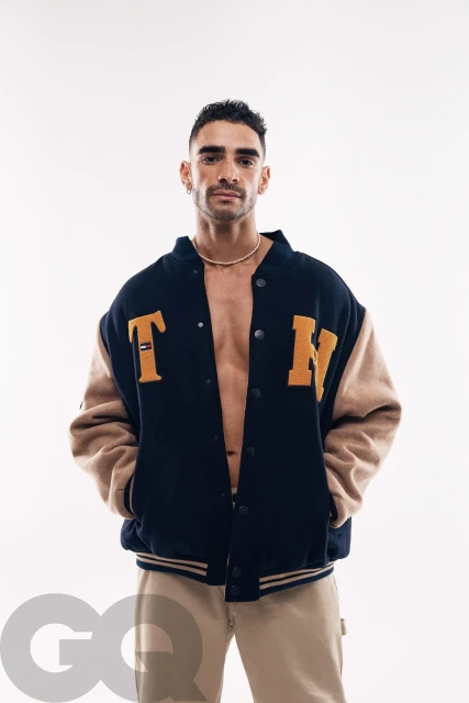 Portrait of a fit, light-skinned male wearing a varsity jacket with open front, neutral pants, and gold chain against a white backdrop.
