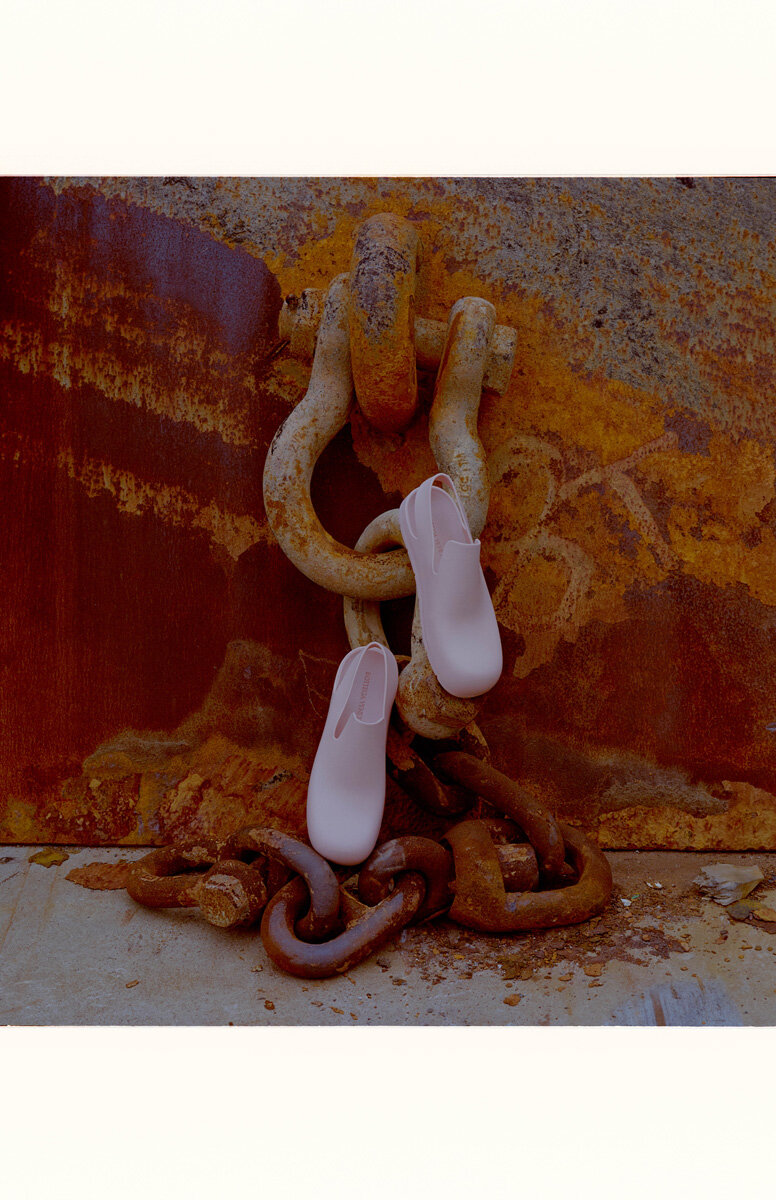 Two light pink shoes draped on a heavily rusted metal chain and U-bolt against a rust-covered steel wall and concrete base indoors.