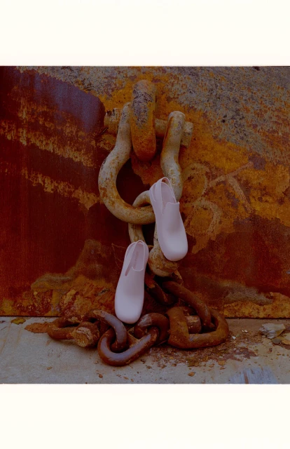 Two light pink shoes draped on a heavily rusted metal chain and U-bolt against a rust-covered steel wall and concrete base indoors.