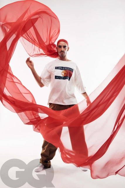 Man wearing a white Timberland t-shirt, brown pants, and a red headscarf, posing in front of a white background with a flowing red fabric.