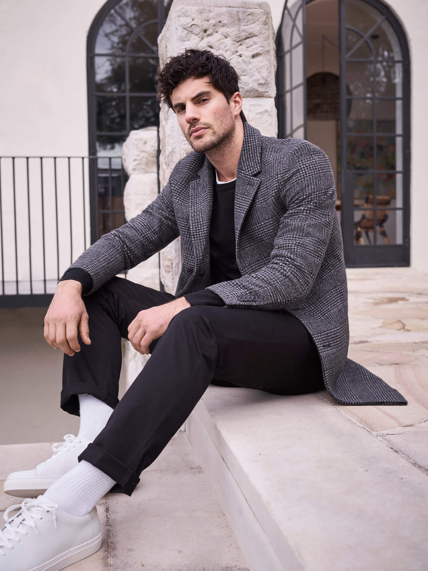 Man sits on steps in casual fashion wear. He's wearing a grey coat, black trousers, white socks and shoes, and a black sweater.