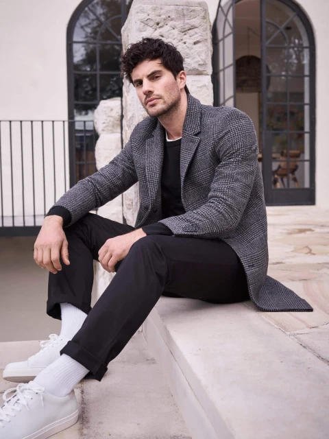 Man sits on steps in casual fashion wear. He's wearing a grey coat, black trousers, white socks and shoes, and a black sweater.