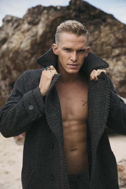 Man with short blonde hair in a herringbone coat stands outside on a sandy beach, posing with an open jacket and rocky background.