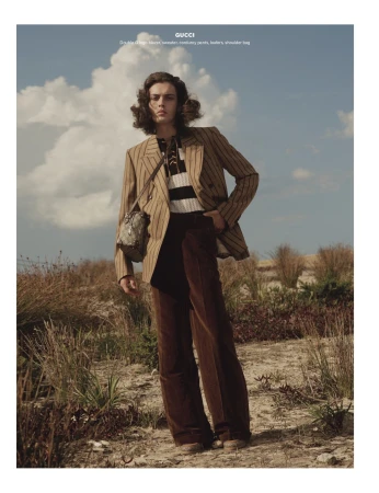Model with long wavy hair wearing striped blazer, striped sweater, and corduroy pants outdoors by coast