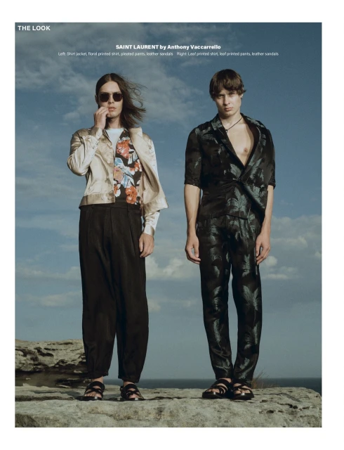 Two male fashion models in Saint Laurent apparel pose outdoors on a rocky outcrop with a sky and water background, showcasing editorial style.
