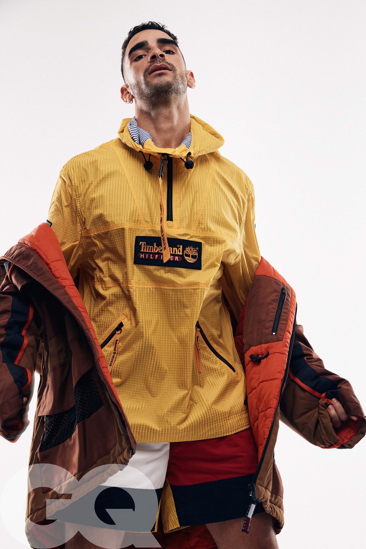 Man poses in a yellow hooded jacket with a Timberland logo, shorts, and jacket with orange lining against a white backdrop.