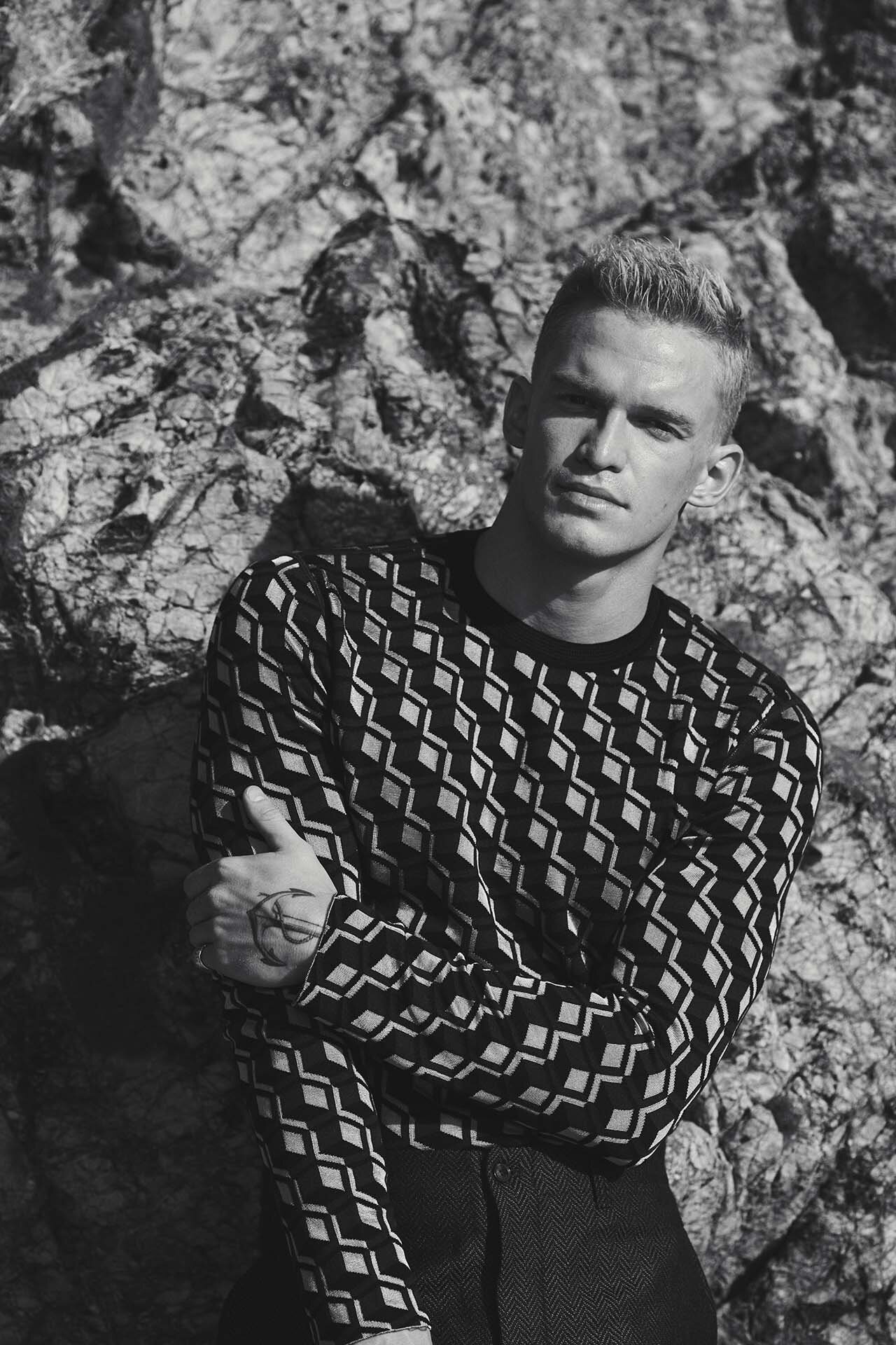 Monochrome portrait of a young man wearing a patterned long-sleeve top against a rocky natural background