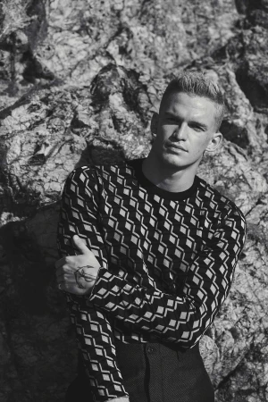 Monochrome portrait of a young man wearing a patterned long-sleeve top against a rocky natural background