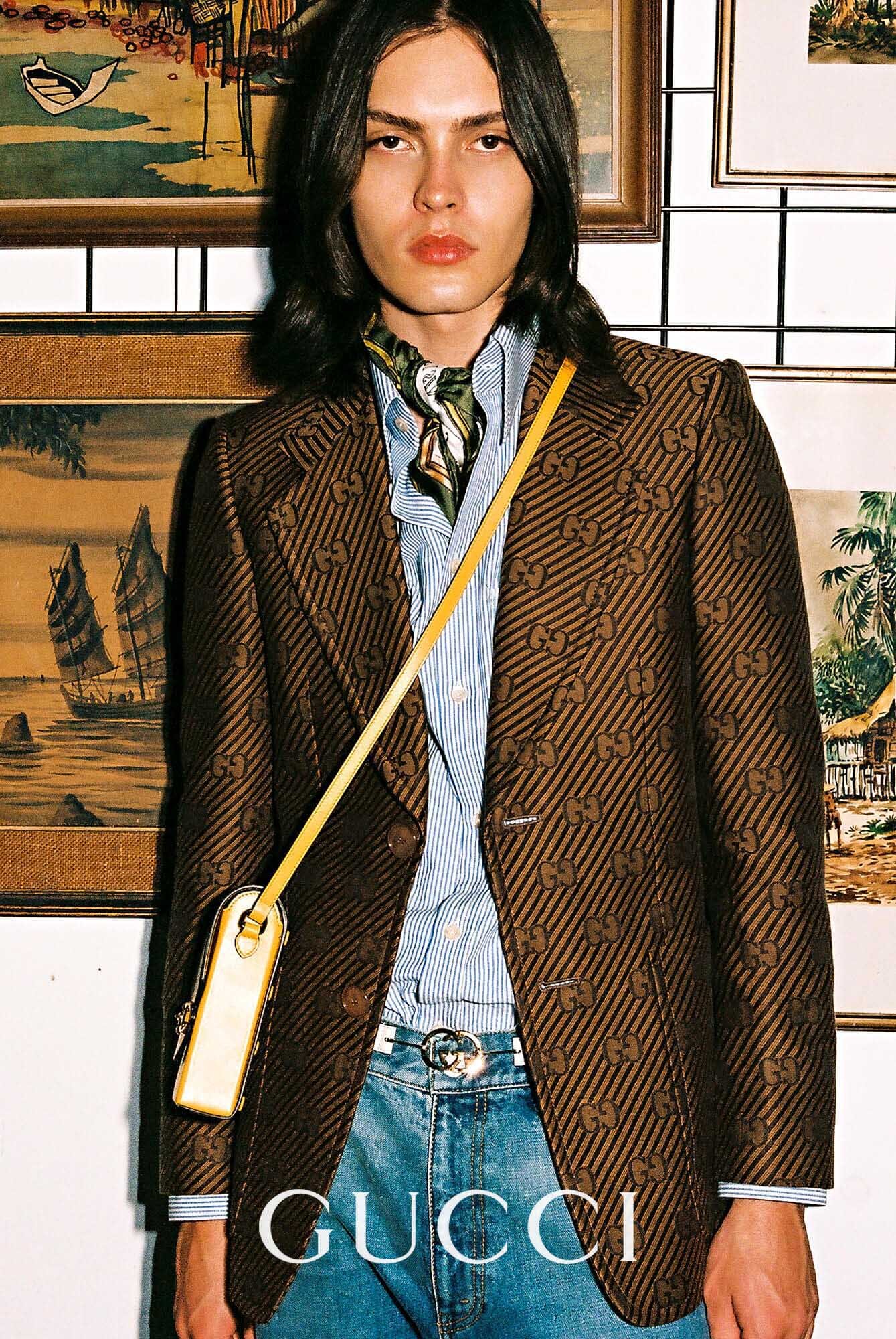 Young adult male poses in Gucci blazer, blue jeans, and neck scarf, standing indoors against a wall with framed art pieces.