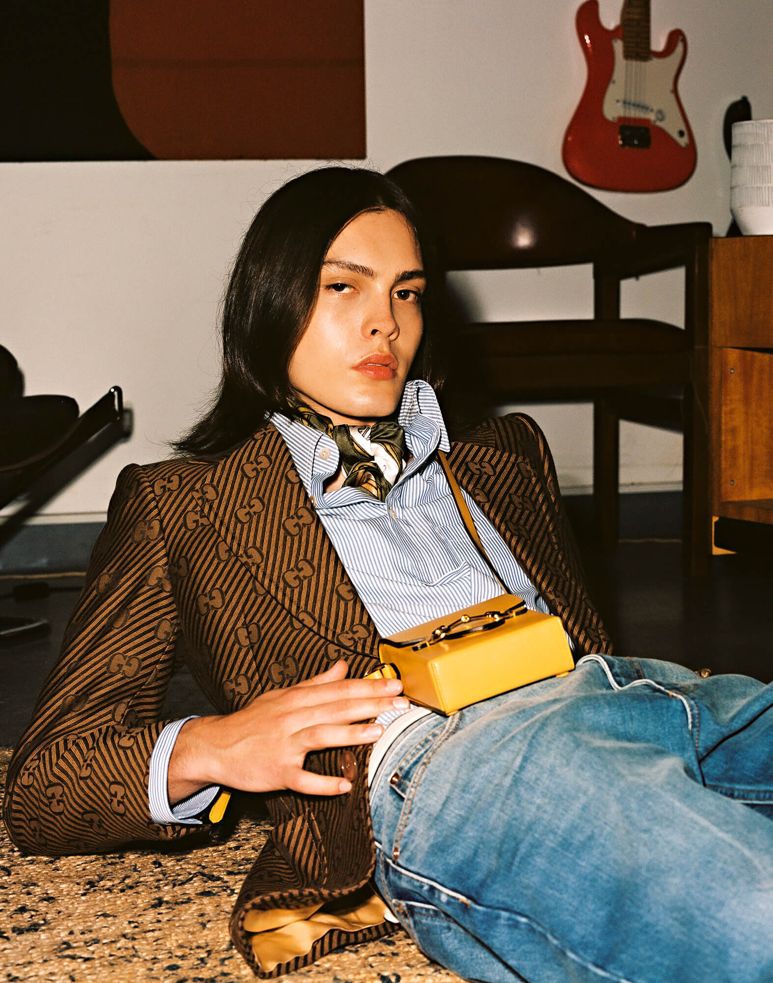 Person lying on carpet with a brown patterned blazer, blue jeans, neck scarf, and a small yellow purse on their lap in a room.