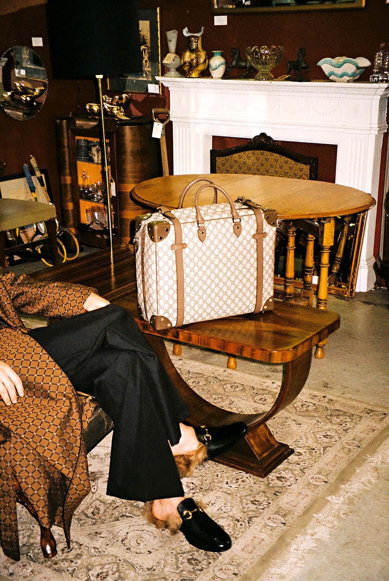 Tan patterned satchel purse sits on a wooden table beside a person in patterned jacket, black pants, and black fur-lined loafers.