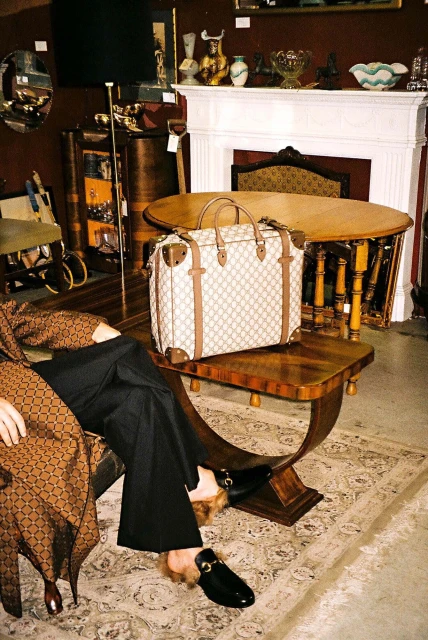Tan patterned satchel purse sits on a wooden table beside a person in patterned jacket, black pants, and black fur-lined loafers.