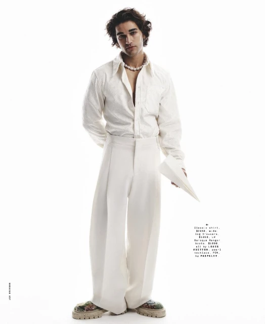 Young man in an unbuttoned white shirt and wide-leg trousers poses against a bright white background in a studio environment.