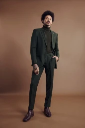 Man with dark curly hair wearing a dark green suit, dark turtleneck, and brown leather boots against a solid warm brown background