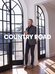 A mature man in a grey blazer and dark trousers stands smiling indoors against arched windows, styled by Grant Pearce.