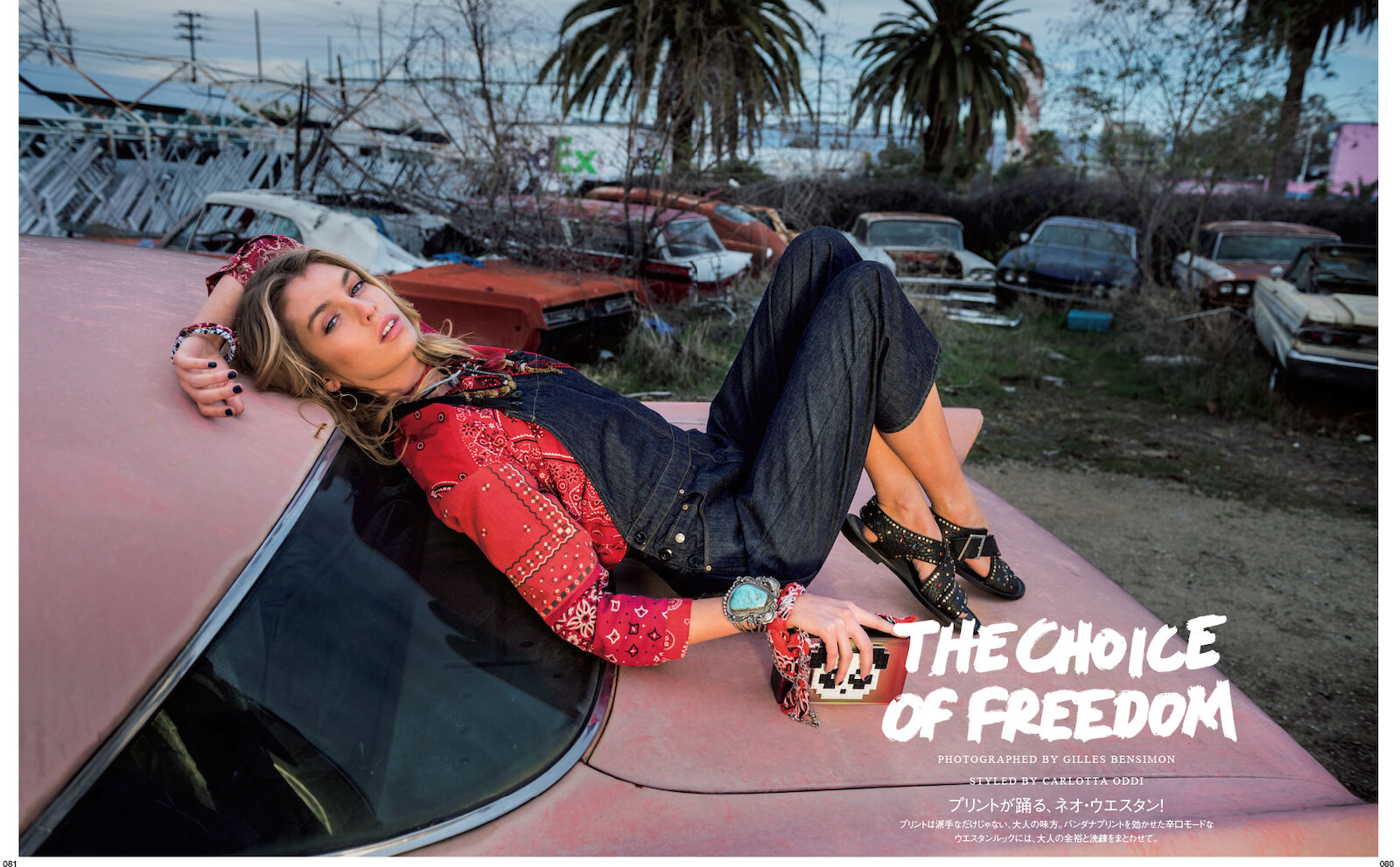 Adult female model in denim overalls and red bandana-print shirt poses on a vintage car in a junkyard under an overcast sky. 