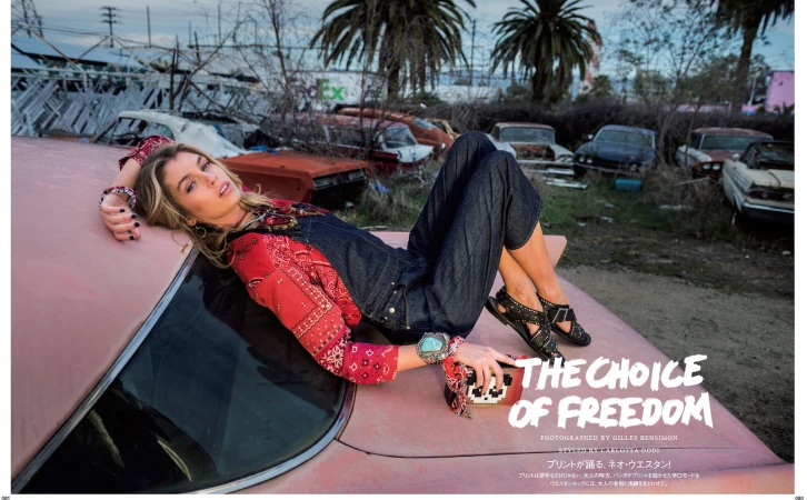Adult female model in denim overalls and red bandana-print shirt poses on a vintage car in a junkyard under an overcast sky. 