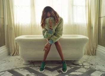 Woman in fuzzy green patterned sweater and green sneakers sitting on a white bathtub in a marble bathroom.