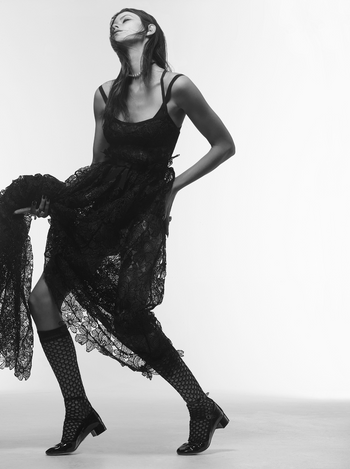 A slender woman wearing a flowing lace dress and dotted hosiery poses in a dramatic stance against a stark white backdrop in black and white.