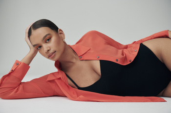 A woman with dark hair lies on a white surface in a black bodysuit and salmon-colored shirt, shot in a studio with soft lighting.