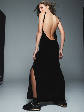 A slender woman with curly hair, seen from behind, wears a low-back black dress and athletic shoes in a fashion photo studio.