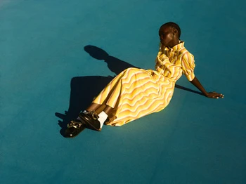 Woman in a patterned yellow dress and black shoes sits on a bright turquoise background, casting a sharp shadow.