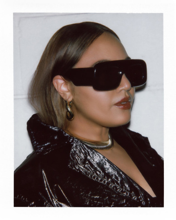 A portrait of a woman wearing oversized sunglasses and a shiny black jacket against a white wall with a retro, vintage feel.
