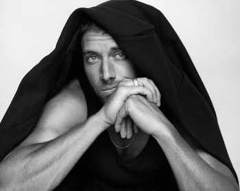 Monochrome portrait of a man with a dark jacket draped over his head, resting his chin on his hands against a plain light background