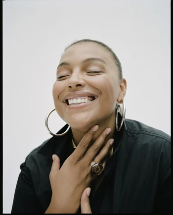 Portrait of a smiling woman with eyes closed wearing large silver hoop earrings and a dark collared shirt against a white wall.