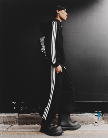 Man in black Adidas tracksuit with white stripes and heavy lug-sole boots walks against dark wall and sidewalk.