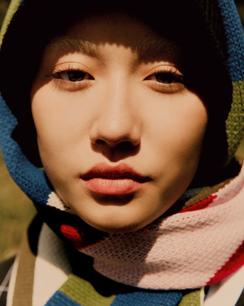Close-up portrait of a young adult woman with fair skin, fringed hair, and a multicolored knit hood against an outdoor background.