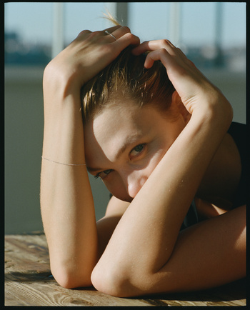 Eye-level shot of a blonde adult woman leaning on a wood table indoors with hands in her hair. Natural light from a window.