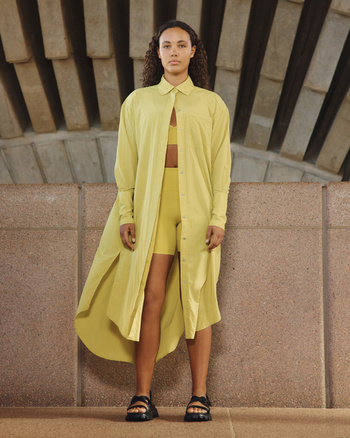 Woman in a yellow shirt dress and matching workout set poses against a textured concrete wall, showcasing a casual yet stylish outfit.
