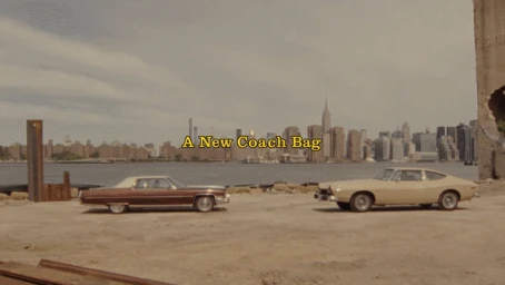 Two vintage cars parked on dirt ground with a city skyline and waterfront in the background