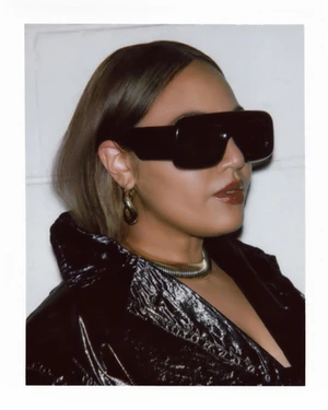 Portrait of a woman wearing large black sunglasses, shiny black jacket, gold necklace and hoop earrings against a white background