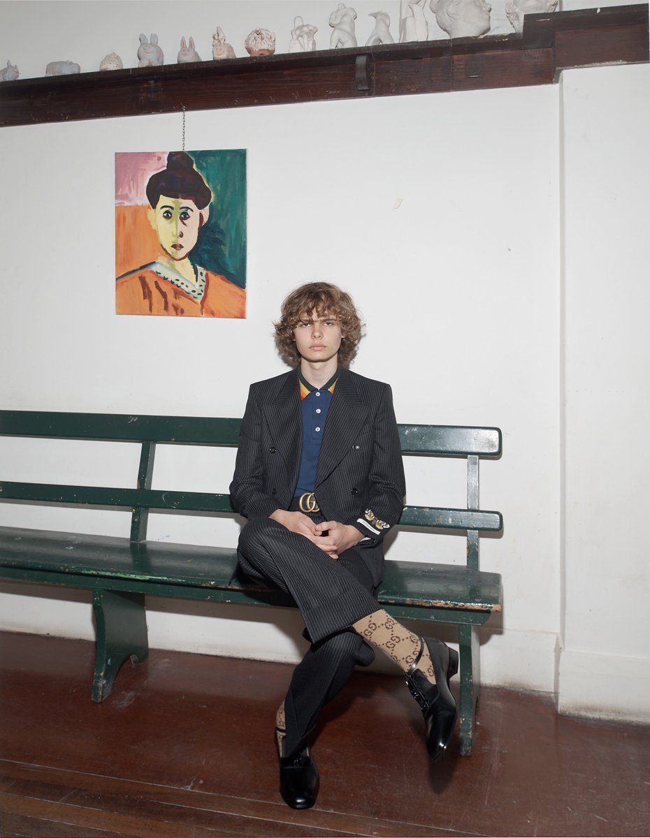 Man with curly hair sitting on a green bench inside, wearing a black blazer, blue polo shirt, patterned pants, socks, and black shoes.
