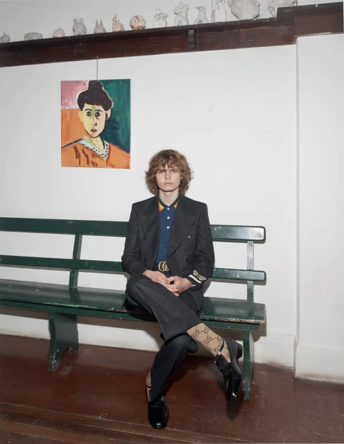 Man with curly hair sitting on a green bench inside, wearing a black blazer, blue polo shirt, patterned pants, socks, and black shoes.